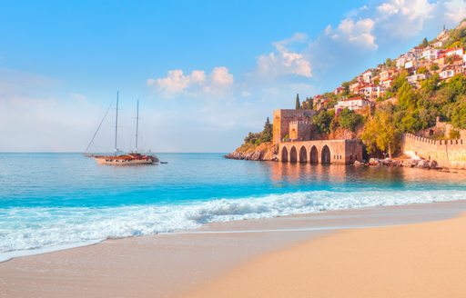 A gulet anchored in blue waters opposite a steep hill nestled with houses leading down to a medieval fortress