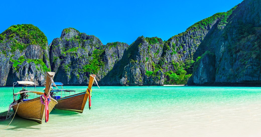 Long-tailed boats are pulled up onto a golden beach lapped by green waters with a limestone rocky outcrop in the distance