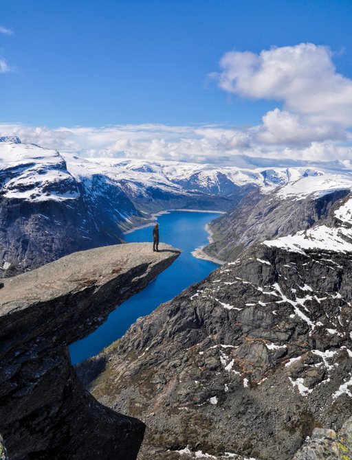 A lone figure stands at the end of Pulpit Rock in Norway and surveys the majestic landscape of snow-capped mountain peaks